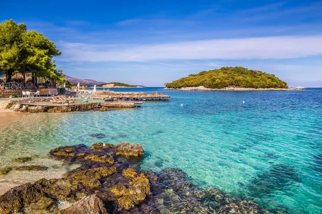 Felsige Küste mit klarem, türkisfarbenem Wasser, das zu einem kleinen Strand mit einem Außencafé und einer grünen Insel in der Ferne führt.