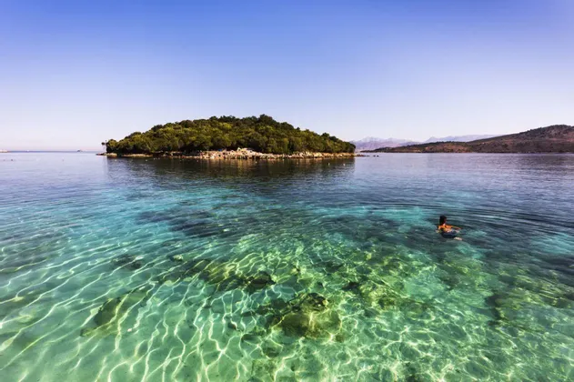 Una persona nada en aguas cristalinas de color turquesa cerca de una pequeña isla arbolada bajo un cielo azul brillante.