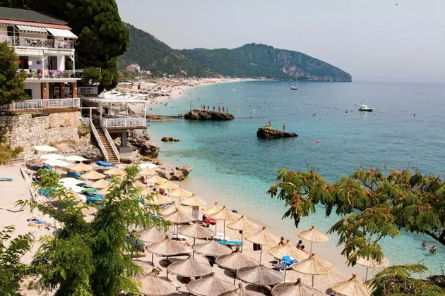 Ein erhöhter Blick auf einen belebten Strand mit vielen Strohschirmen, klarem türkisfarbenem Wasser und einer bewaldeten Bergküste.