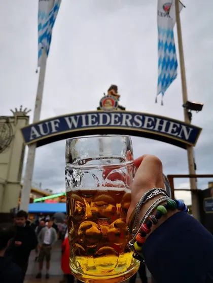 Una mano con braccialetti regge un grande boccale di birra di vetro di fronte a un cartello con la scritta 'AUF WIEDERSEHEN' durante un festival.