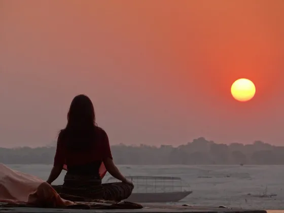 Una persona vista di spalle è seduta in posizione meditativa, mentre osserva il sole tramontare su un fiume nebbioso.