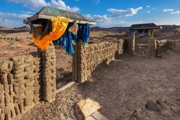 Un portail et un mur en pisé décorés de tissus jaunes et bleus vifs, dans un vaste paysage aride sous un ciel partiellement nuageux.