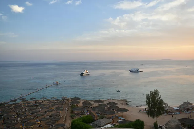 Una vista dall'alto di una spiaggia sabbiosa con ombrelloni di paglia e sedie a sdraio, con diversi yacht sul mare calmo al tramonto.