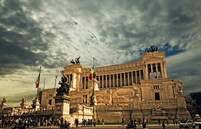 Un grande monumento classico con colonne, statue e bandiere italiane si erge sotto un cielo scuro e nuvoloso mentre le persone passano.