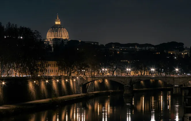 Una cupola illuminata si erge su uno skyline notturno della città, con un ponte di pietra e lampioni che si riflettono nel fiume sottostante.