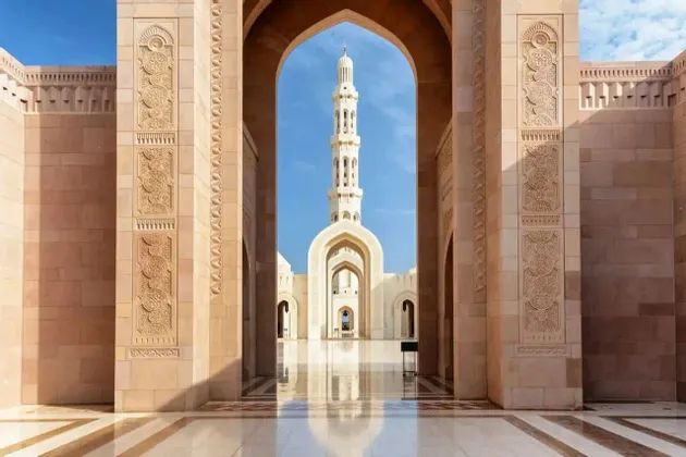 Ein hohes Minarett einer Moschee ist durch einen prächtigen Steinbogen von einem Innenhof mit einem polierten, spiegelnden Boden zu sehen.