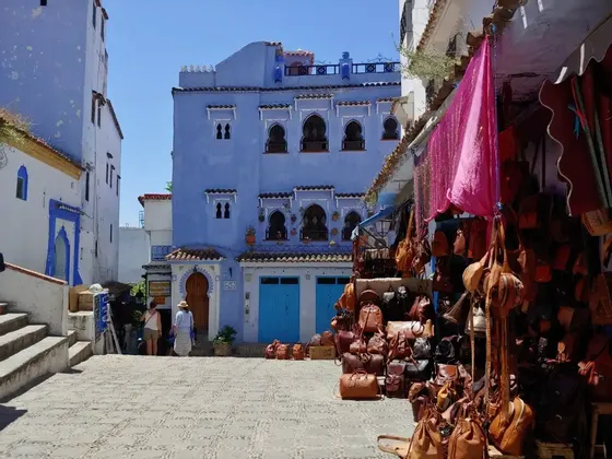Auf einem Kopfsteinpflasterplatz vor einem großen, blauen Gebäude bietet ein Marktstand Ledertaschen zum Verkauf an.