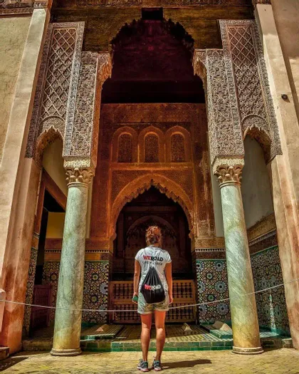 Une femme portant un t-shirt et un sac à dos WeRoad se tient debout, regardant une grande arche ornée de sculptures détaillées et de mosaïques en carreaux.