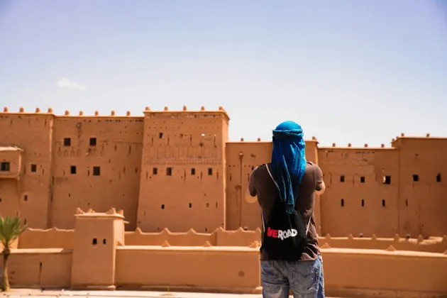 Eine Person von hinten, die ein blaues Kopftuch und einen WeRoad-Rucksack trägt, blickt auf eine große, sandfarbene Kasbah unter klarem Himmel.