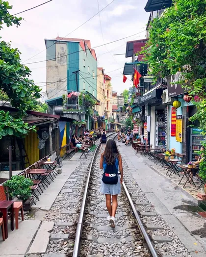 Une femme avec un sac à dos WeRoad marche le long d'une voie ferrée traversant une rue étroite bordée de cafés.