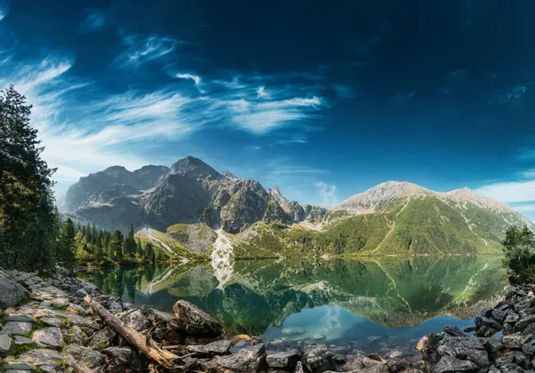 Un lac alpin aux eaux claires reflète parfaitement une chaîne de montagnes rocheuses sous un ciel d'un bleu profond avec des nuages légers, une rive de galets au premier plan.