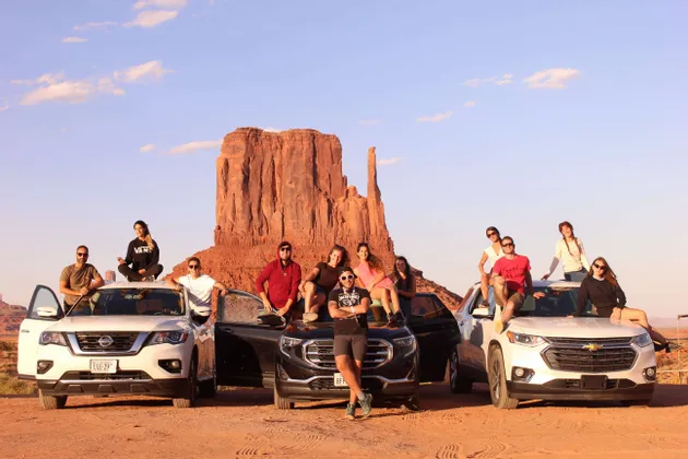 Un viaggio di gruppo WeRoad con persone in posa su tre auto davanti a grandi formazioni rocciose rosse in un paesaggio desertico.