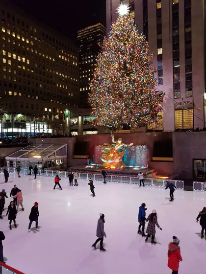 Personas patinando sobre hielo de noche en una pista al aire libre frente a un gran árbol de Navidad iluminado y altos edificios de la ciudad.