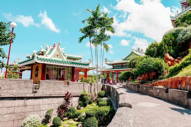 Templi sontuosi con tetti di tegole verdi circondati da giardini lussureggianti, palme e sentieri in pietra sotto un cielo blu.