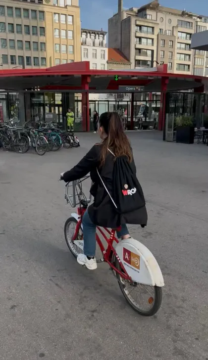 Una donna vista di schiena, con uno zaino WeRoad nero, pedala su una bicicletta rossa attraverso una piazza cittadina pavimentata.