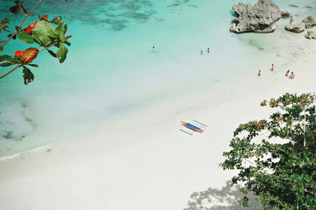 Veduta aerea di una spiaggia di sabbia bianca con persone che nuotano in acque cristalline turchesi e una canoa a bilanciere sulla riva.