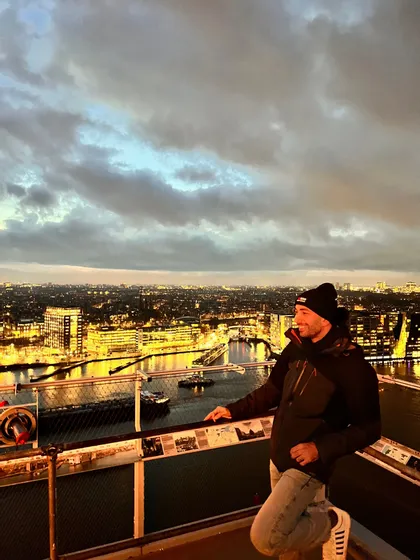 Un uomo con berretto e giacca si trova su una piattaforma panoramica, che si affaccia su un paesaggio urbano illuminato e un fiume al tramonto.