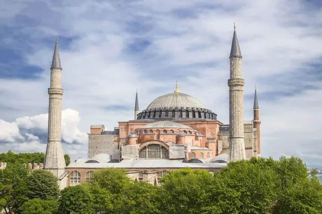 Eine große Moschee mit zentraler Kuppel und vier Minaretten erhebt sich über grünen Bäumen vor einem teils bewölkten Himmel.