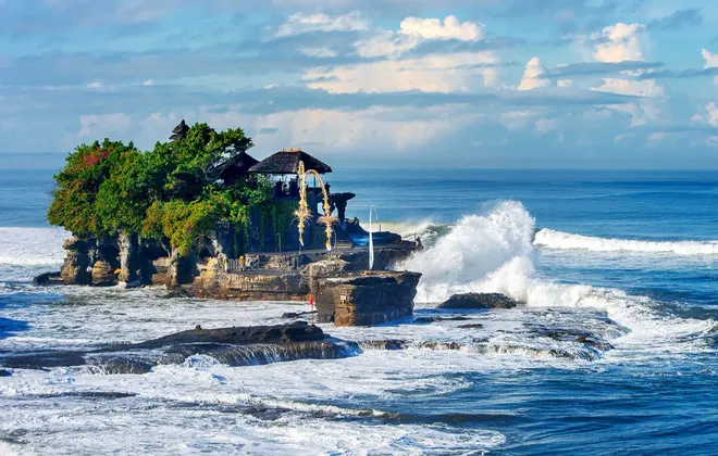Un templo se asienta en una pequeña isla rocosa cubierta de árboles, con grandes olas del océano rompiendo contra la orilla bajo un cielo parcialmente nublado.