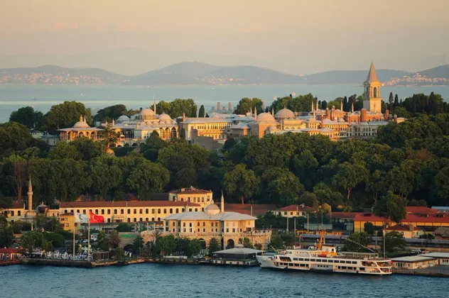 Ein großer Palast mit Kuppeln und Türmen, eingebettet in Bäume an einem Ufer, mit Blick auf das Wasser, wo bei Sonnenuntergang eine Fähre anlegt.