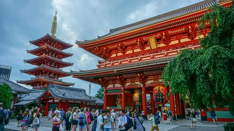 Una multitud de personas se reúne en un patio frente a una gran puerta roja de templo y una pagoda de cinco pisos bajo un cielo nublado.