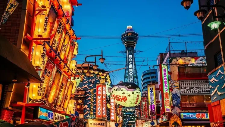 Letreros y faroles iluminados, incluido un gran pez globo, bordean una calle de la ciudad con una torre visible al fondo contra un cielo azul crepuscular.