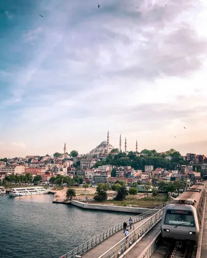 Vogelperspektive: Ein Zug überquert eine Brücke über Wasser, im Hintergrund eine historische Stadt mit markanter Moschee.