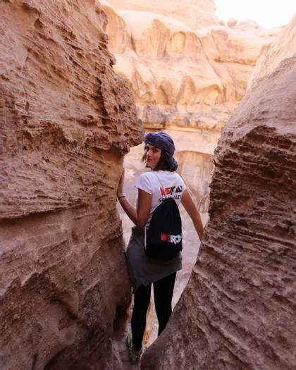 Una mujer con una camiseta de WeRoad y una mochila mira hacia atrás y sonríe mientras camina por un estrecho cañón de roca.