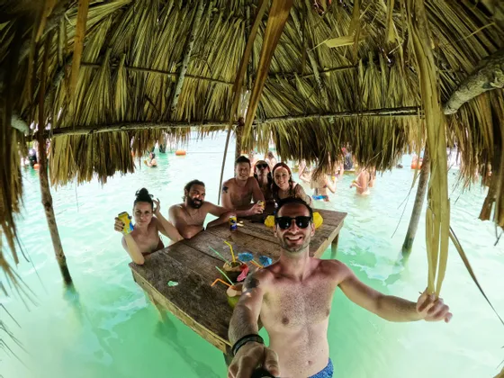 Eine WeRoad-Gruppe macht ein Selfie beim Trinken an einem Tisch, der teilweise im türkisfarbenen Wasser unter einem Strohdach steht.