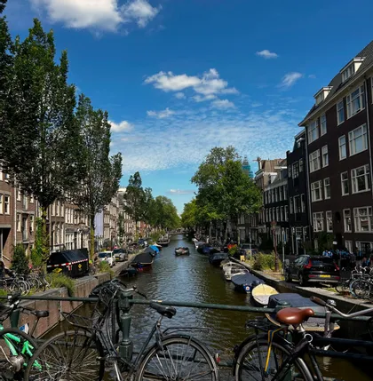 Vélos garés sur un pont surplombant un canal de ville bordé de hauts immeubles, d'arbres et de bateaux sous un ciel bleu.