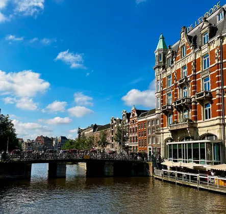 Un pont bondé de vélos traverse un canal de la ville, avec des bâtiments historiques en briques rouges longeant les quais sous un ciel bleu clair.