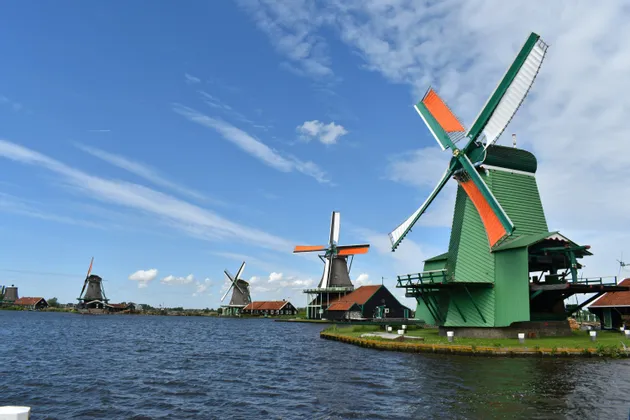 Traditionelle Windmühlen, eine davon leuchtend grün gestrichen, stehen am grasbewachsenen Ufer eines Wasserwegs unter blauem Himmel.