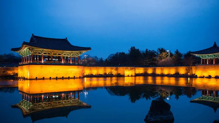 Un tradizionale padiglione e muro dell'Asia orientale, illuminato da luci gialle, che si riflette su un laghetto calmo al crepuscolo.