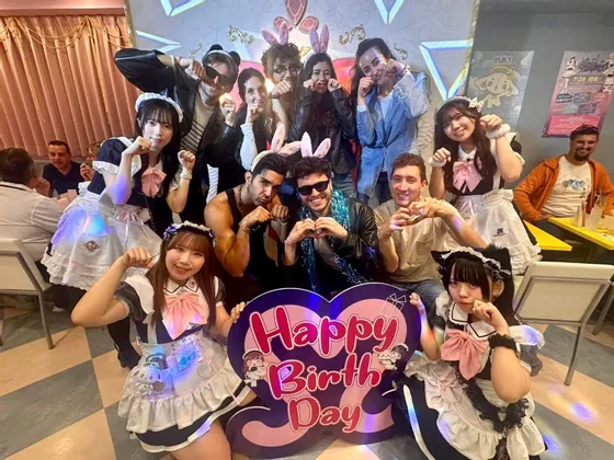 Un gruppo WeRoad in viaggio posa con donne in abiti da cameriera in un caffè a tema, reggendo un grande cartello 'Buon Compleanno'.