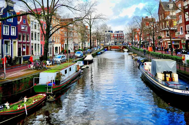 Des bateaux sont amarrés le long d'un canal urbain bordé de bâtiments colorés, avec des gens qui se promènent dans la rue adjacente sous un ciel partiellement nuageux.