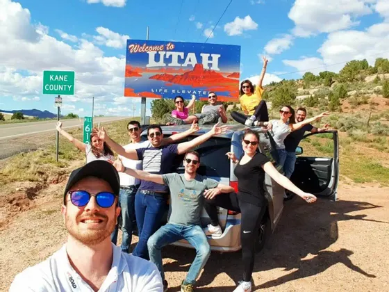 Un gruppo WeRoad in viaggio si scatta un selfie sorridente con la propria auto davanti al cartello 'Welcome to Utah'.