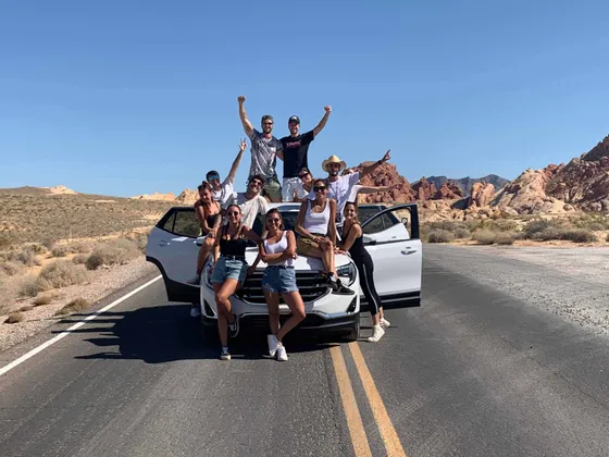Un viaggio di gruppo WeRoad in posa per una foto su e intorno a un SUV bianco su una strada in un deserto con formazioni rocciose rosse.