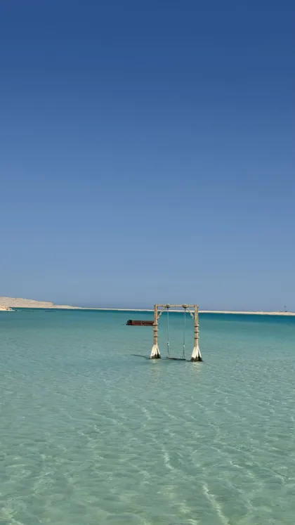 Un'altalena di legno si erge in acque poco profonde e limpide di colore turchese sotto un cielo di un azzurro intenso, con una costa sabbiosa in lontananza.