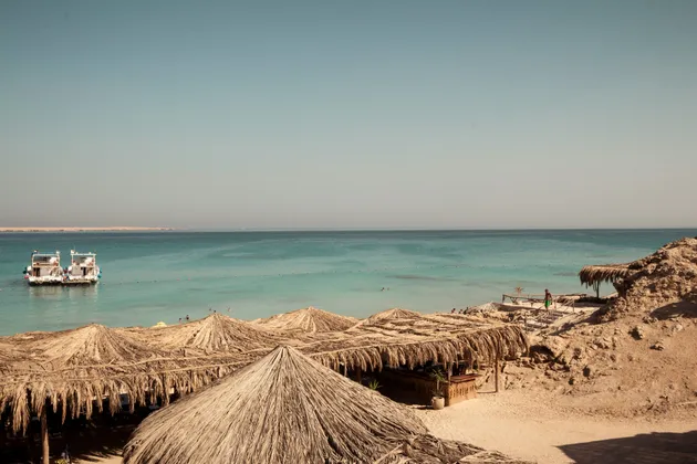 Una vista dall'alto di una spiaggia sabbiosa con ombrelloni dal tetto di paglia, che si affaccia su due barche ormeggiate in acqua turchese sotto un cielo sereno.