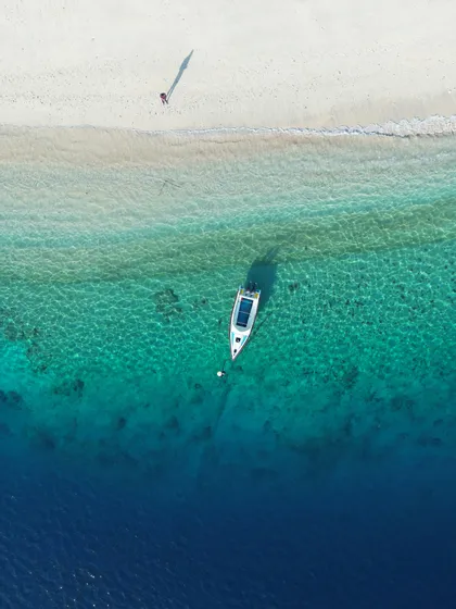 Vista aerea di una barca ancorata in acqua turchese accanto a una spiaggia di sabbia bianca con una persona che cammina lungo la riva.