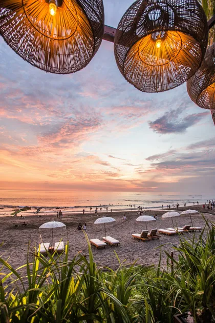 Paralumi intrecciati pendono sopra una spiaggia sabbiosa con lettini e ombrelloni mentre le persone si godono un tramonto colorato sull'oceano.