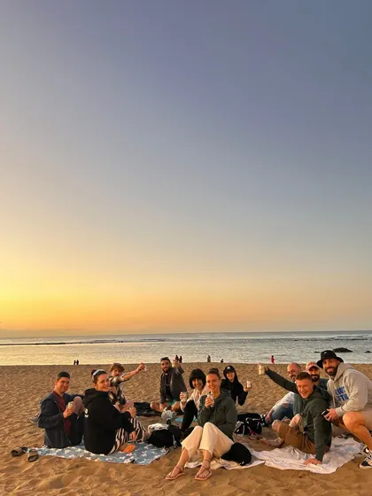 Eine WeRoad-Gruppe sitzt auf Decken an einem Sandstrand und genießt Getränke während eines goldenen Sonnenuntergangs über dem Meer.