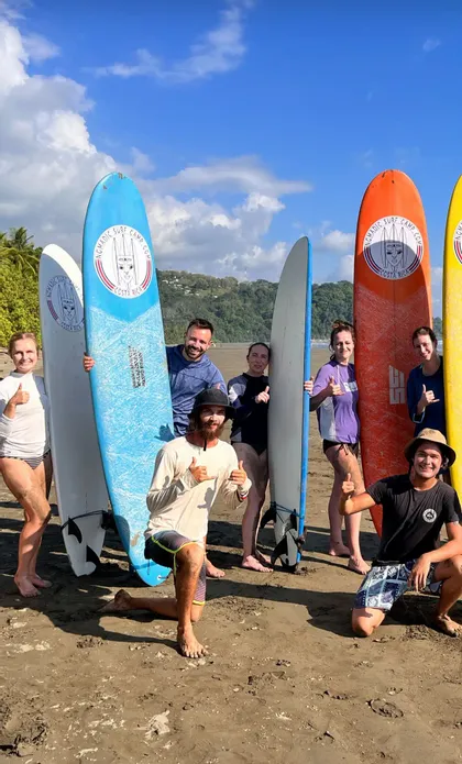 Eine WeRoad Gruppenreise posiert mit bunten Surfbrettern an einem Sandstrand unter blauem Himmel.