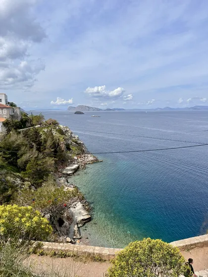 Una vista dall'alto di una costa rocciosa con alberi verdi, una piccola caletta con acqua turchese e isole lontane sotto un cielo nuvoloso.