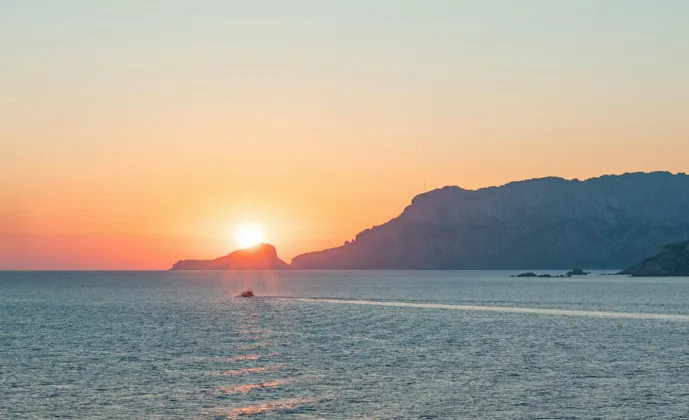 Una piccola barca attraversa il mare al tramonto, lasciando una scia mentre il sole scende dietro una costa in controluce.