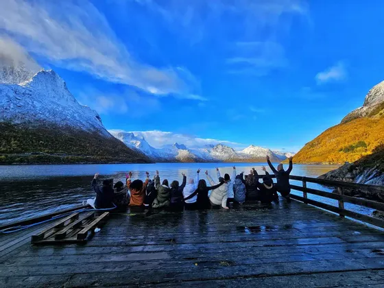 Un gruppo WeRoad è seduto su un molo di legno bagnato con le braccia alzate, di fronte a un fiordo e a montagne innevate.
