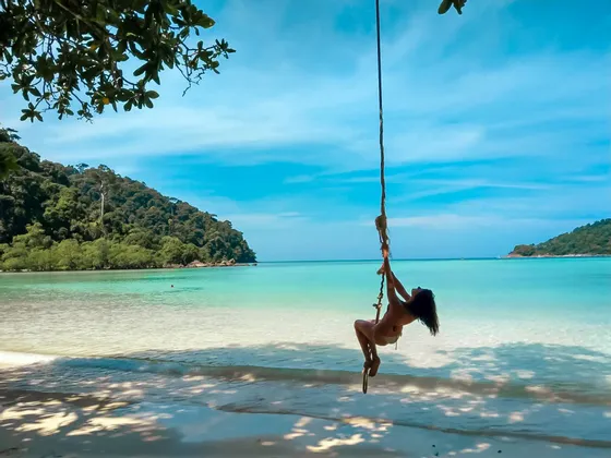 Une femme en bikini sur une balançoire en corde au-dessus d'une plage tropicale avec sable blanc et eau turquoise claire.