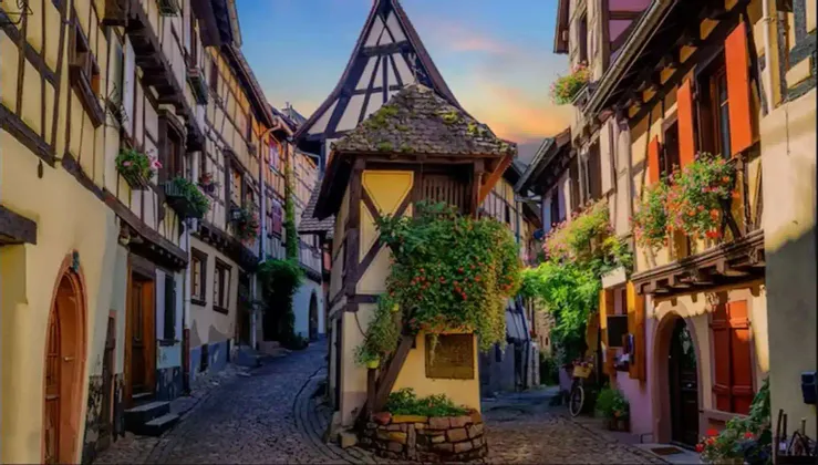 Eine schmale Kopfsteinpflastergasse schlängelt sich zwischen farbenfrohen Fachwerkhäusern, die mit Blumen und Pflanzen geschmückt sind.
