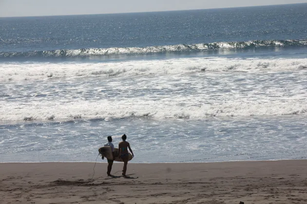 Un uomo e una donna trasportano tavole da surf camminando su una spiaggia sabbiosa verso le onde dell'oceano.