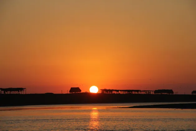 Il sole tramonta all'orizzonte su uno specchio d'acqua, stagliando una costa con diverse piccole capanne di paglia.